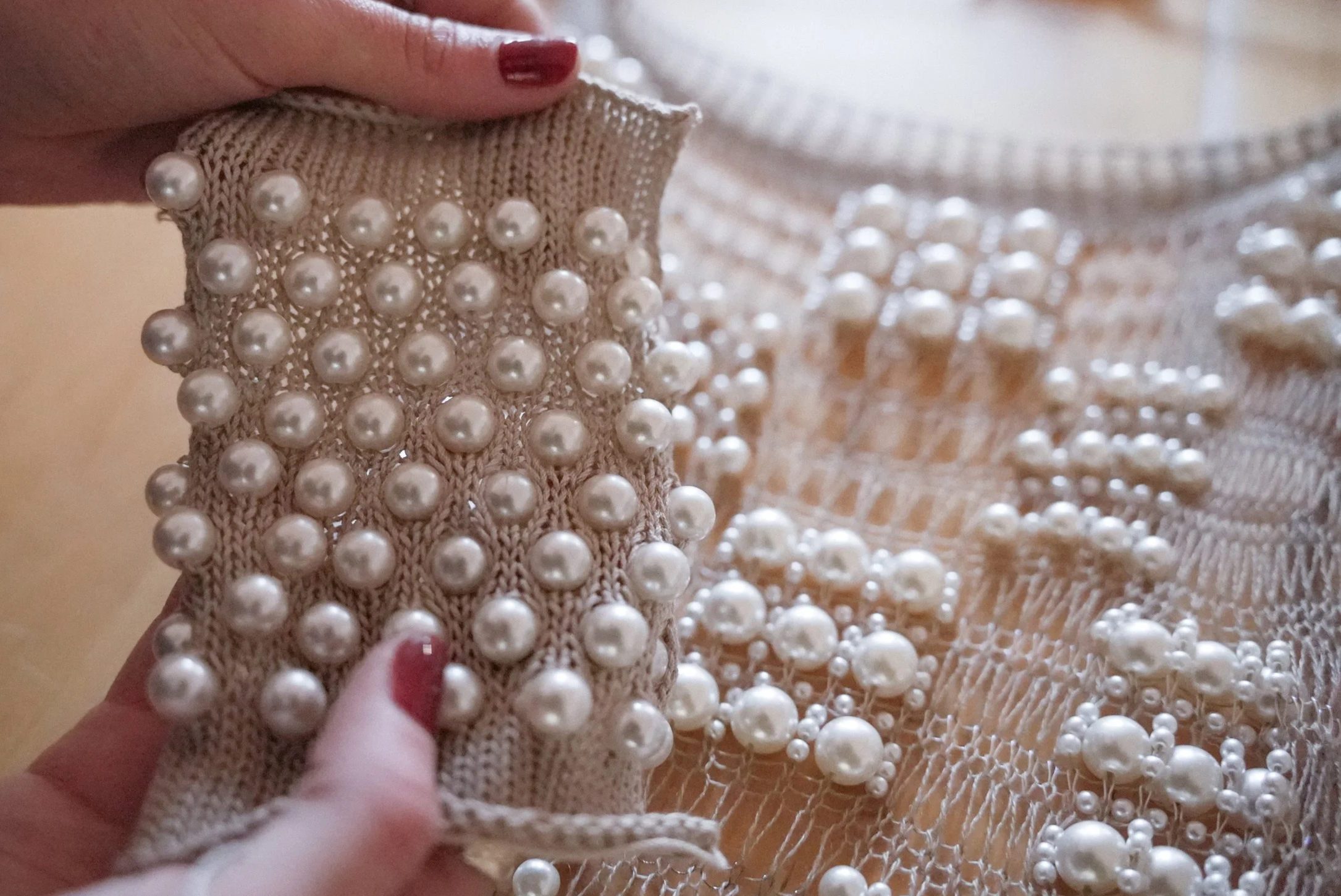 Close-up of a hand holding a knitted fabric with pearl embellishments, with a background of more knitted fabric with pearl details.