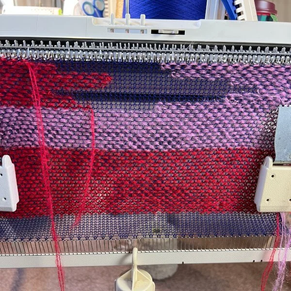 A knitting machine with woven fabric being made with pink and purple yarns being woven into fabric.