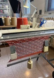 Vintage knitting machine, placed on a work table in a workshop setting with knitting supplies and tools nearby. Fabric with a colourful pattern is hanging down from the machine by a weight.