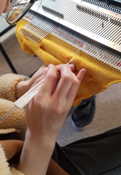 Person working on a knitting machine with a yellow fabric, using a crochet hook to knit or repair the fabric.