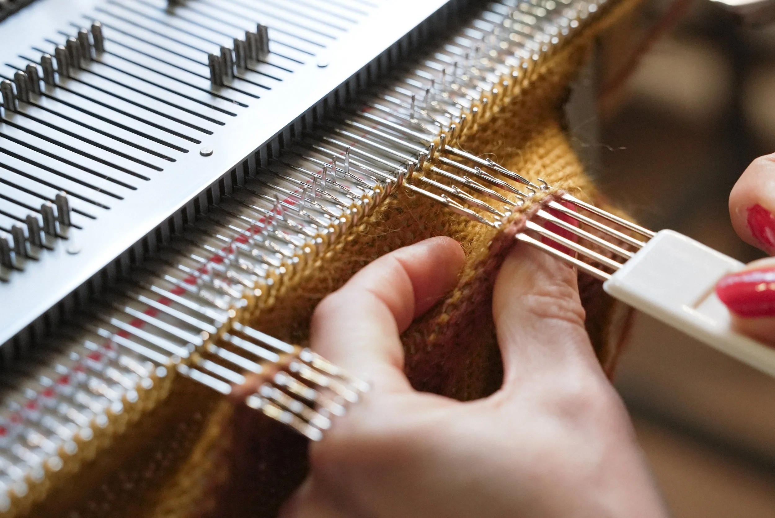 Close-up of a person's hands linking brown fabric on a knitting machine with metal parts.