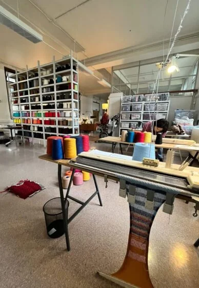 A knitting machine workspace with shelves filled with various supplies and colourful spools of threads and yarn on tables with vintage knitting machines on top.