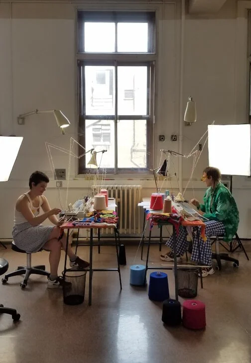 Two young women sitting at separate tables in a bright room, engaged in maching knitting, with bright lights and a large window in the background.