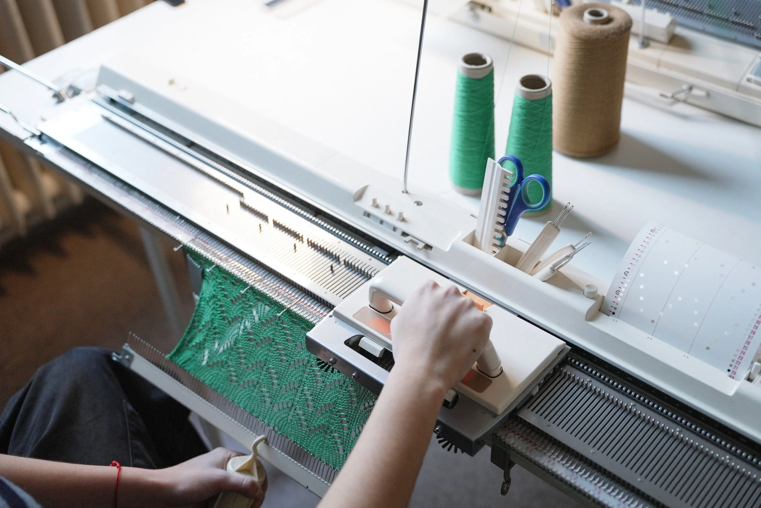Person using a knitting machine to create a green lace knit fabric, with spools of green and beige yarn and knitting tools nearby.