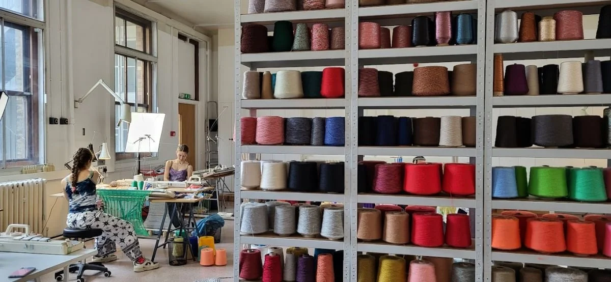 Spools of colorful yarn on shelves in a workshop with two women working at desks near large windows working on vintage knitting machines.
