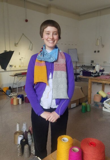 A young woman with short hair smiling, wearing a purple cardigan and a multicoloured knitted scarf, standing in a room with knitting supplies and spools of yarn.