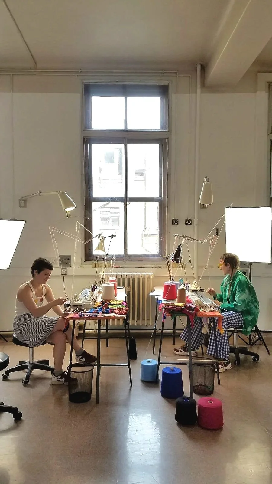Two people sitting at desks, machine-knitting, with bright lighting, a large window in the background, and colourful knits, yarns and materials on the tables. The room is a bright studio workspace, with high ceilings and lots of space.