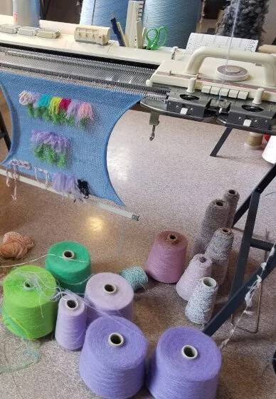 A vintage knitting machine with colourful yarns and spools of thread on the floor.