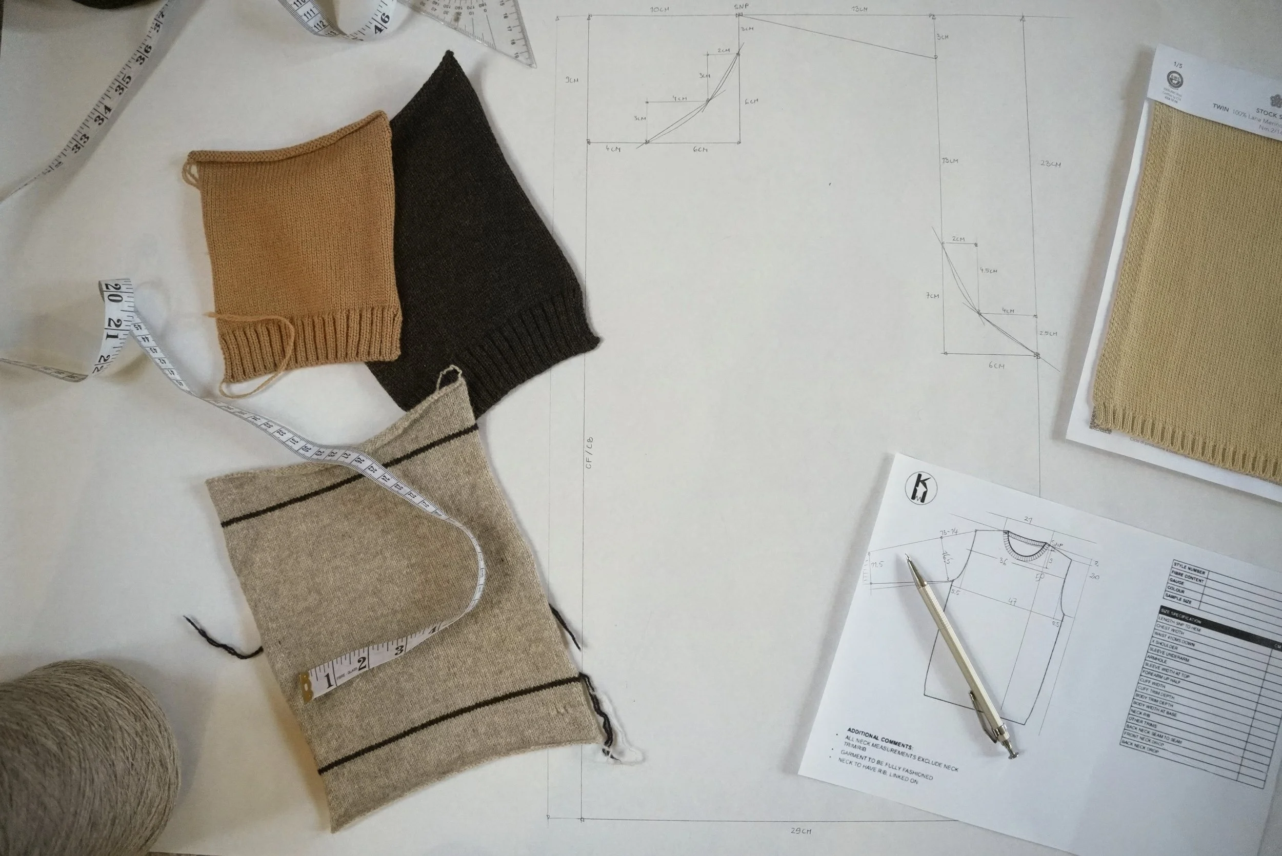 A knit workspace with fabric swatches, measuring tapes, pattern sketches, a pen, and yarn. The fabric swatches include tan, black, cream, and light brown with stripes. The pattern sketches show technical details and measurements for garment design.