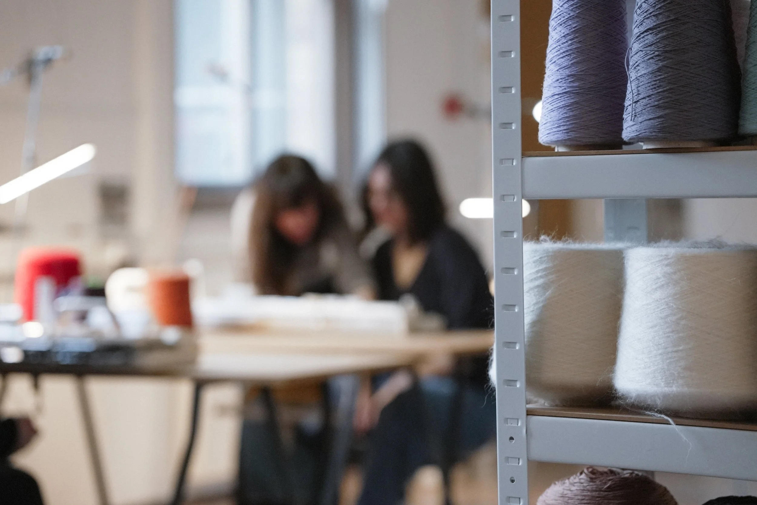 Close-up of shelves with skeins of luxury yarn, with two women blurred in the background working at a vintage knitting machine in a professional textile and knitwear studio.