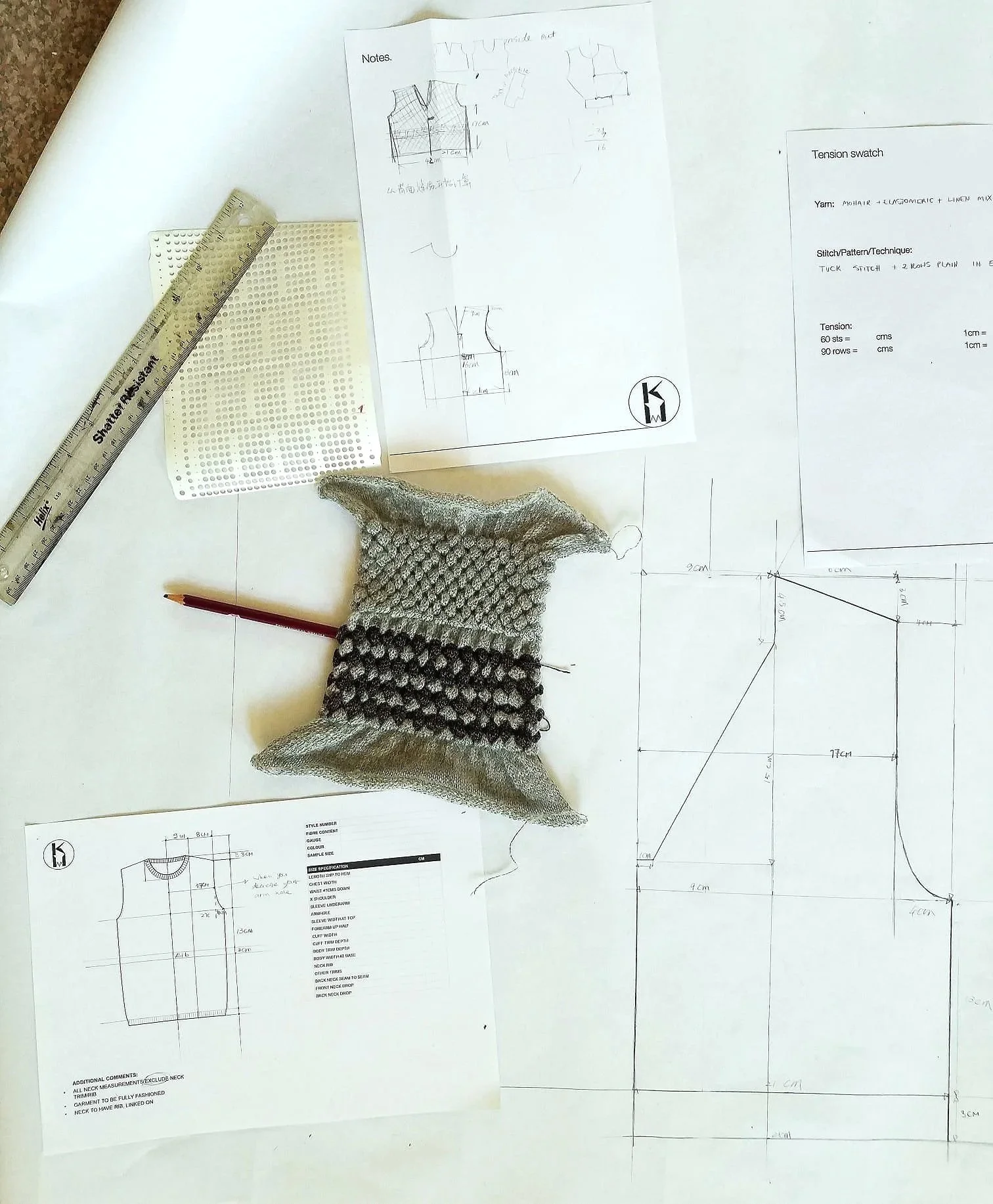 A fashion design workspace with technical sketches, a knitting swatch, a ruler, a pencil, and detailed pattern drafts on large sheets of paper.