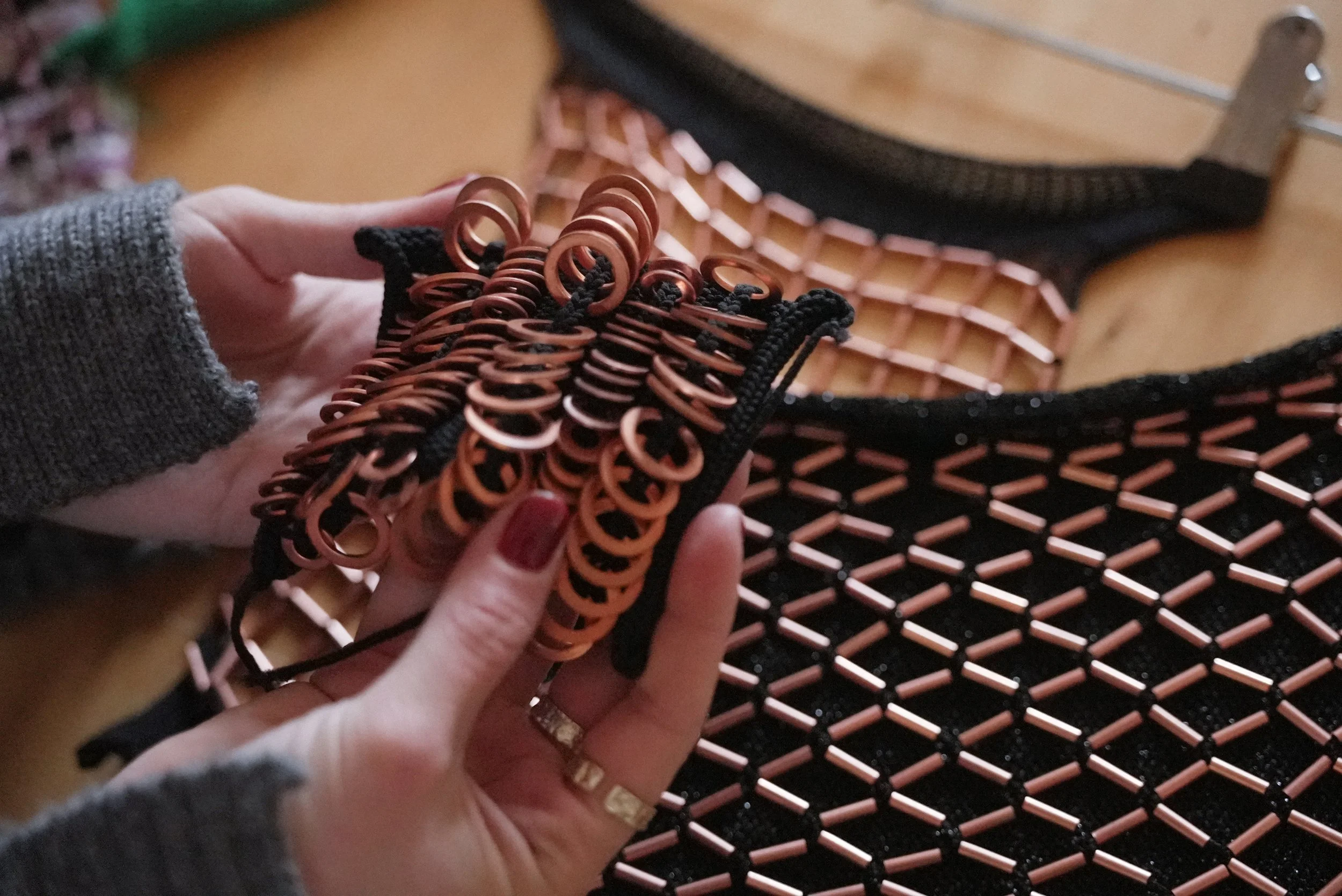 Person holding a small knitted fabric swatch with copper-coloured circular accents, with a wooden surface and black and copper textiles in the background.