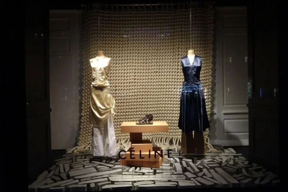 Display window at Saks 5th Avenue, New York, featuring two mannequins in elegant dresses, with a pair of shoes on a small table between them, and the brand name 'CELINE' at the front. In the background is a handmade chunky knit backdrop.