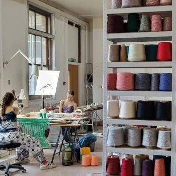Two women working at vintage knitting machines in a bright studio with large windows, colourful spools of thread on shelves, and knitting supplies.