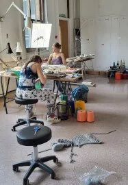Two women working at a large table in a knitwear studio with knit supplies and knitting machines around them, and fabric and yarn lying on the floor.
