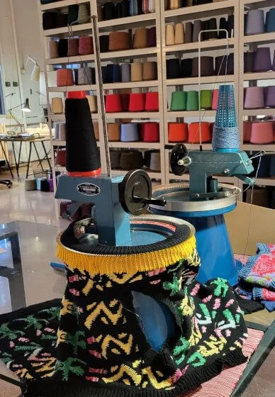 Two knitting machines on a table in a creative knitwear studio, with shelves of colourful yarn in the background.
