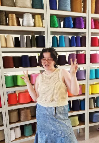 A woman with short dark hair and glasses smiling and making peace signs with both hands, standing in front of shelves of colourful spools of thread. She is wearing a top she has knitted. 
