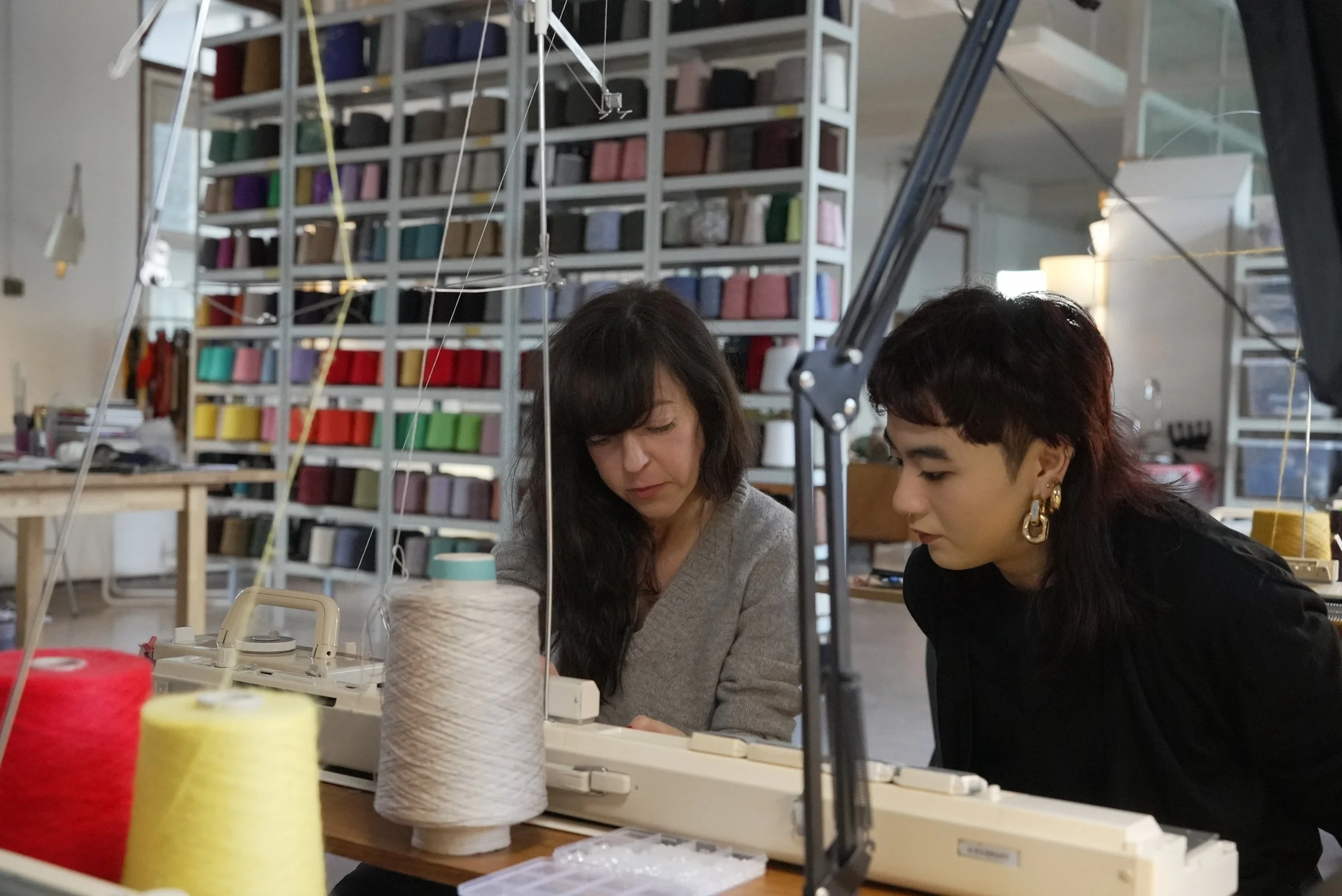 Two women working on a knitting machine in a room with a large shelf of colourful yarn in the background. One woman is teaching the other how to use the vintage knitting machine. Spools of yarn are on the table in front of them.