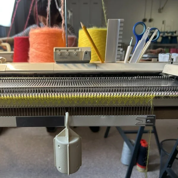 Close-up of a knitting machine with colorful yarn spools, knitting tools, and partially finished fabric in the background.