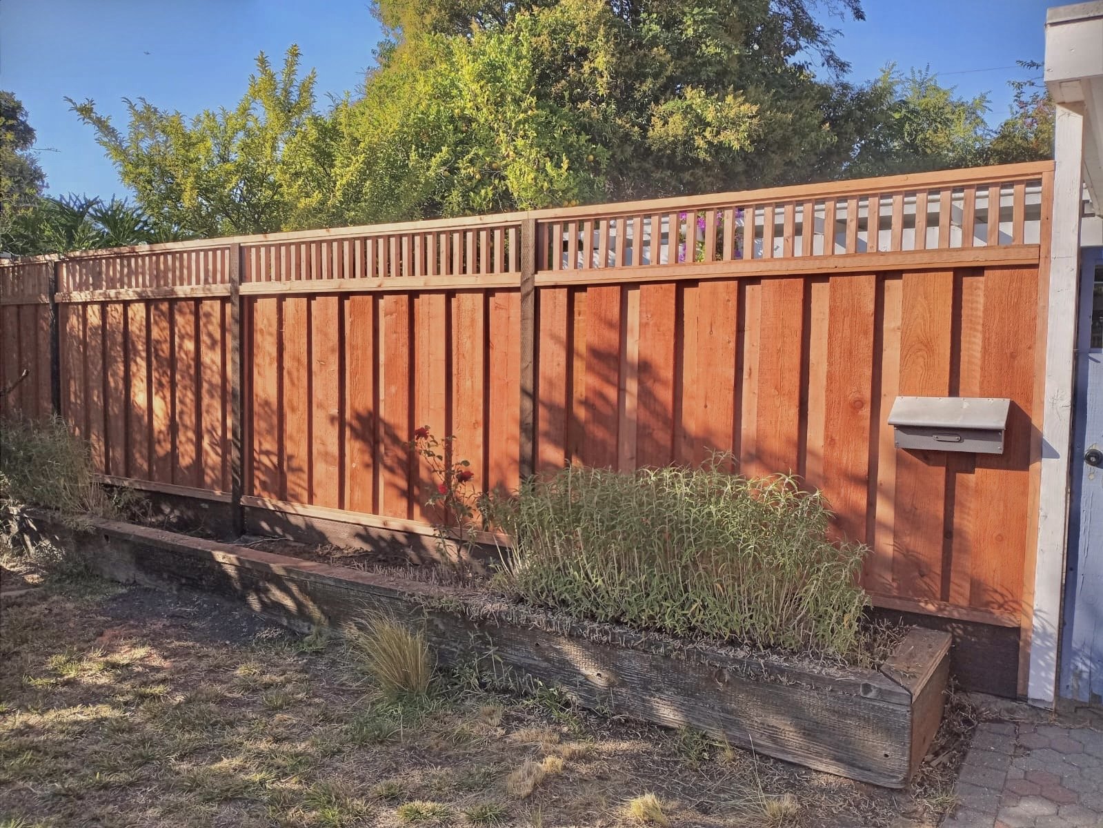 Freshly built red wooden fence with decorative top border surrounding a yard, with a flower bed in front, some plants, and trees in the background under a clear blue sky.