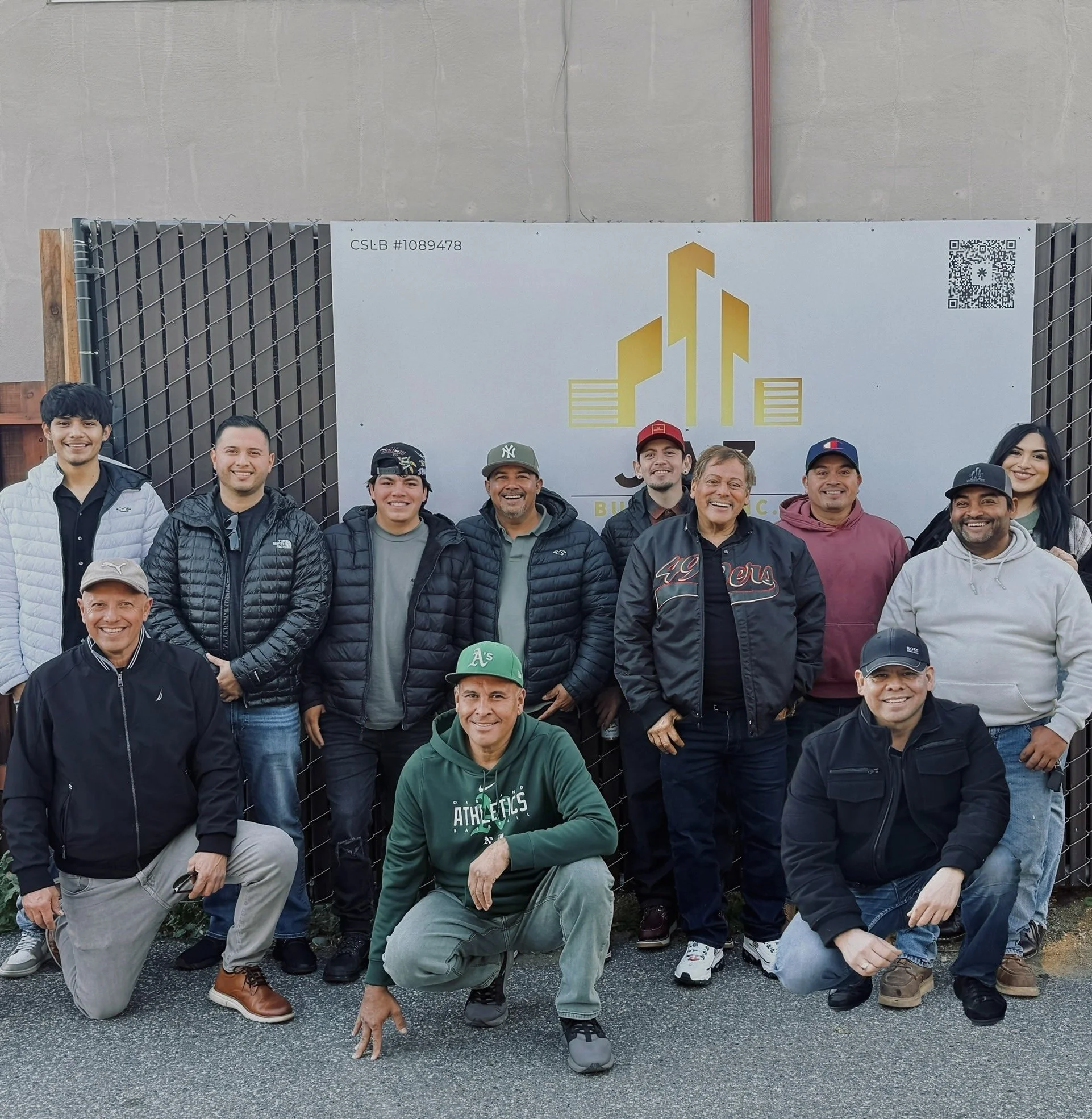 Group of people posing outdoors in front of a construction-themed backdrop with a city skyline logo. The group includes men and women dressed casually, some kneeling, some standing, smiling at the camera.