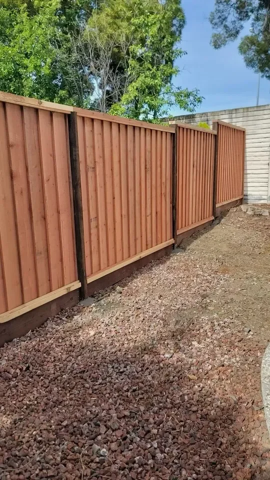 A new wooden privacy fence installed along a yard, with a gravel pathway in front and green trees in the background.
