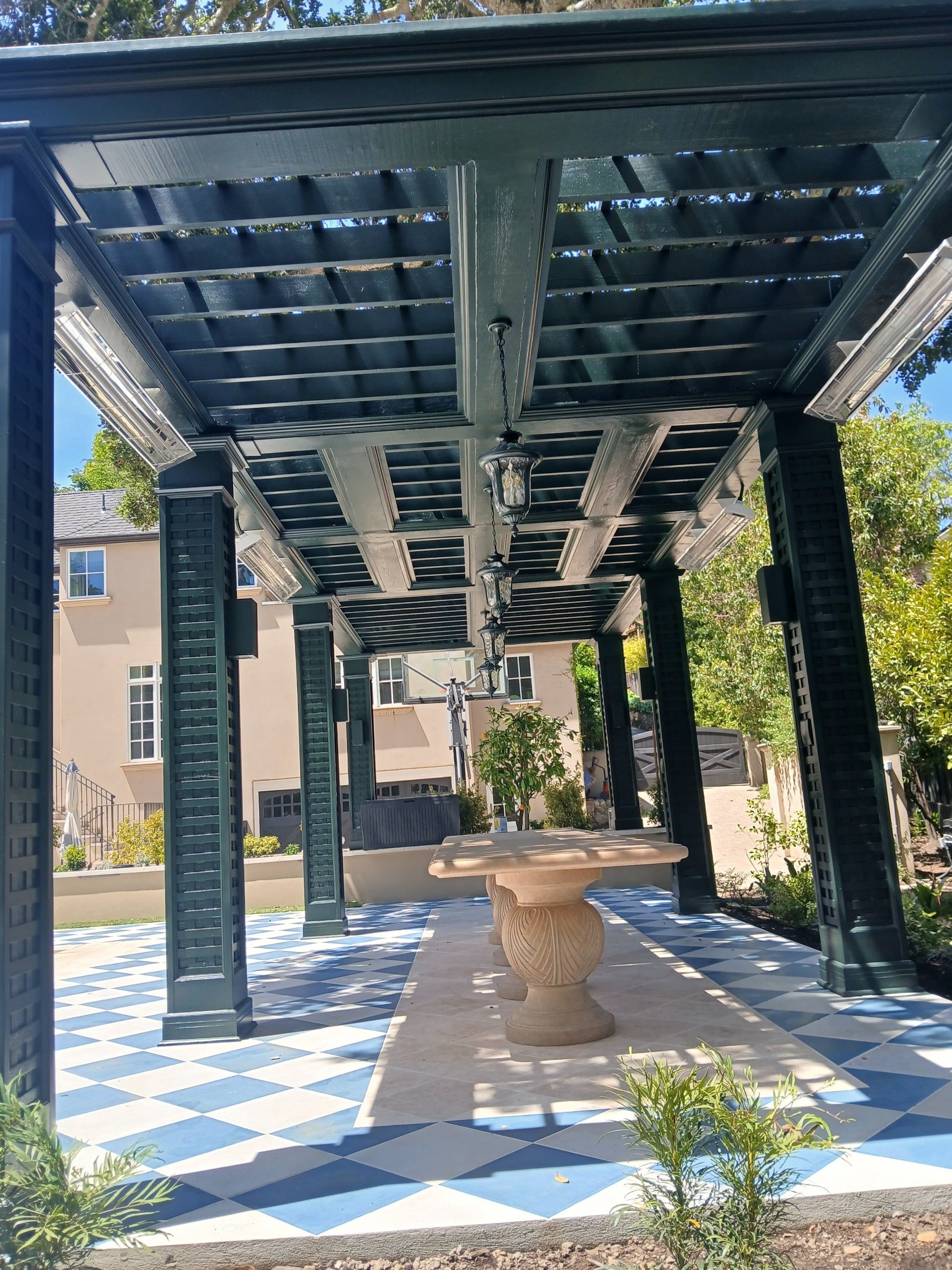 A covered outdoor patio with a checkered blue and white tile floor, a beige stone table with a decorative base, hanging lantern-style lights, and surrounding green trees and plants.