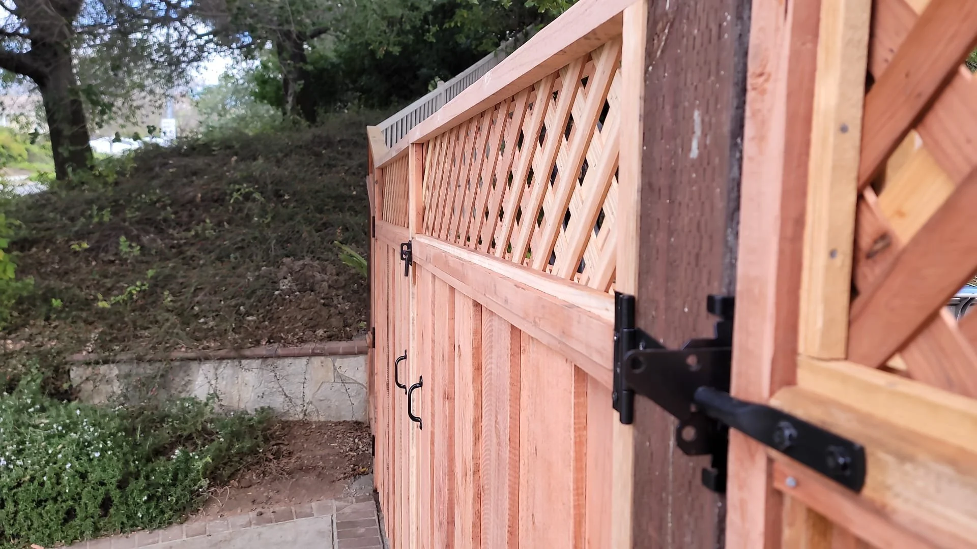 A wooden fence with a gate, featuring a lattice top section and black metal hinges, installed on a brick foundation with surrounding greenery and trees in the background.