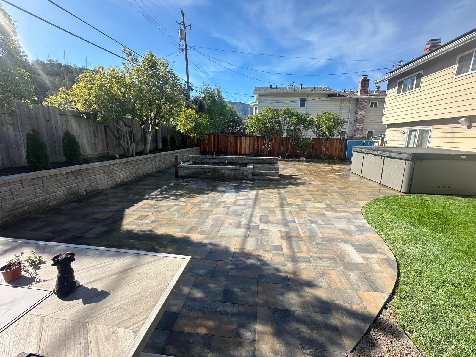 A backyard with a paved stone patio, a small patch of green grass on the right, a hot tub in the far right corner, a raised garden bed in the center, trees along the back fence, and power lines overhead under a blue sky with some clouds.