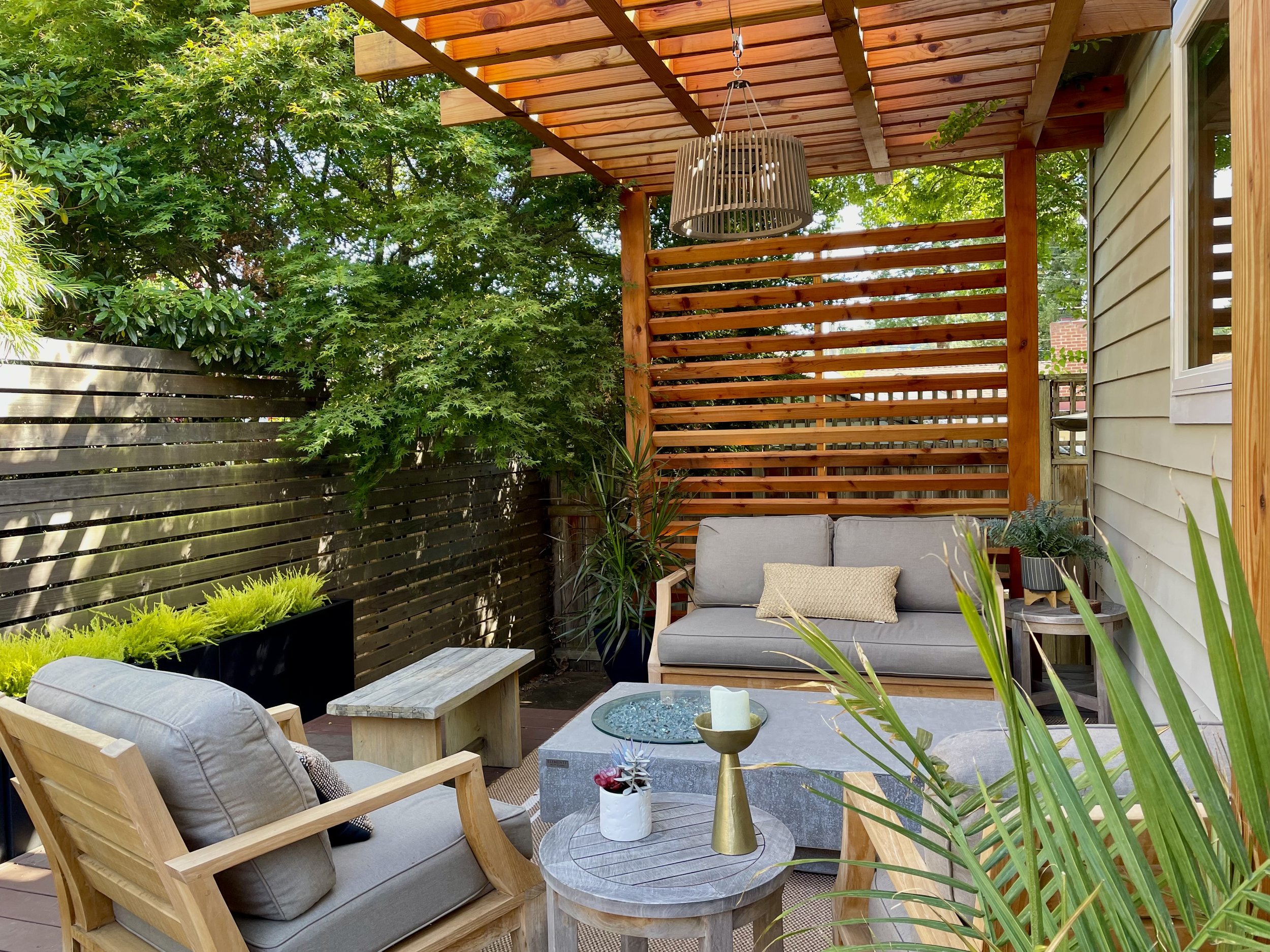 An outdoor patio area with wooden furniture, including a cushioned armchair, a loveseat with cushions, a small wooden bench, and a round side table with a candle and a small decorative plant. There's a wooden pergola overhead with a hanging light fixture. The patio is enclosed by wooden fencing with lush green trees and plants surrounding the space.