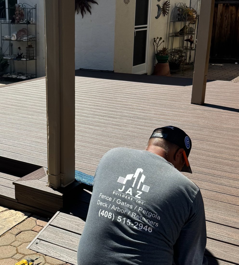 A worker installing or repairing a wooden deck outside a house. The worker is wearing a gray t-shirt with the text 'JAZ Builders, Inc.' and contact information on the back. The scene shows a partially completed deck with some tools visible on the ground.