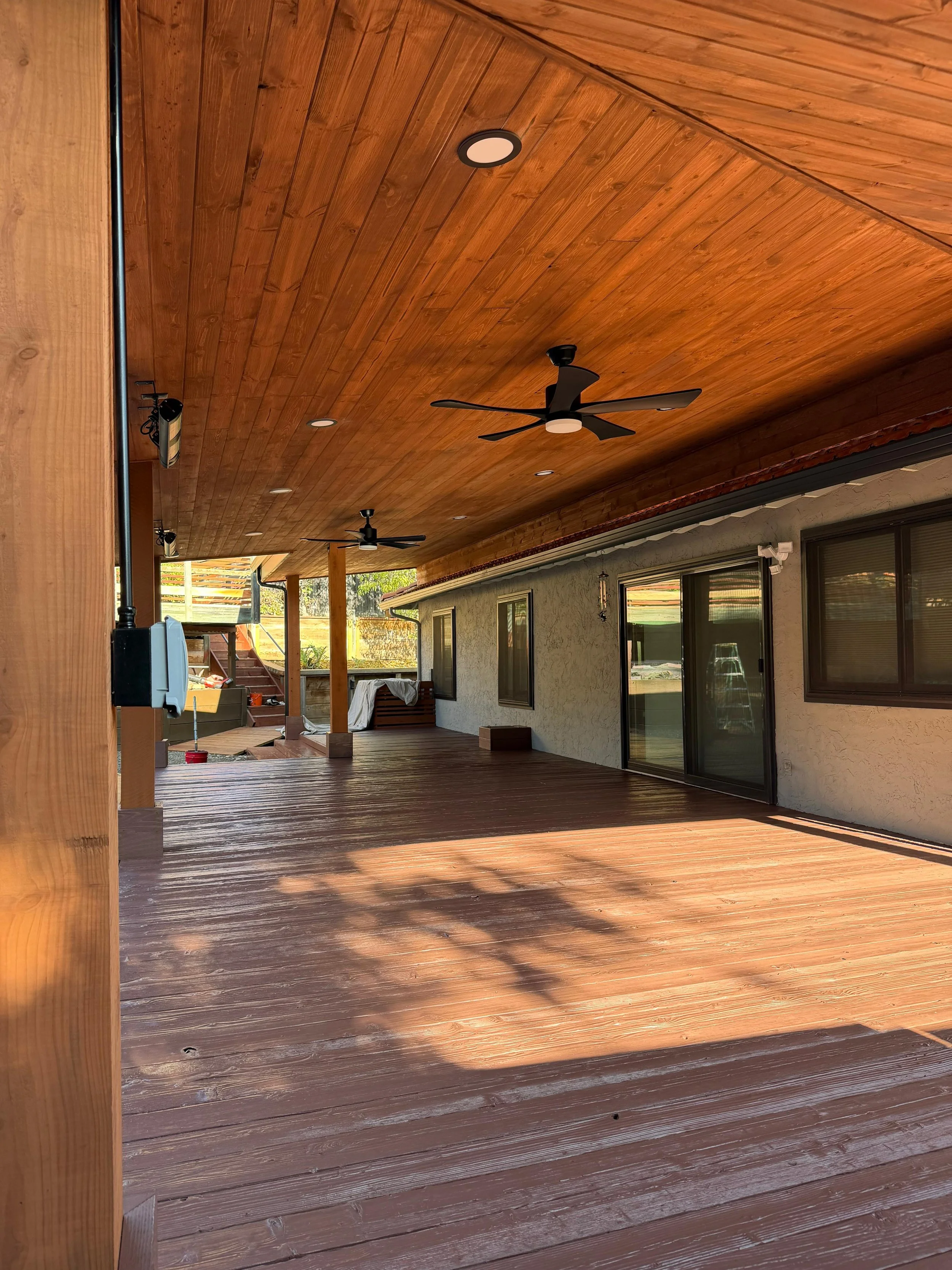 Covered outdoor patio with wooden flooring, a wooden ceiling with recessed lighting and ceiling fans, and sliding glass doors leading into a house.