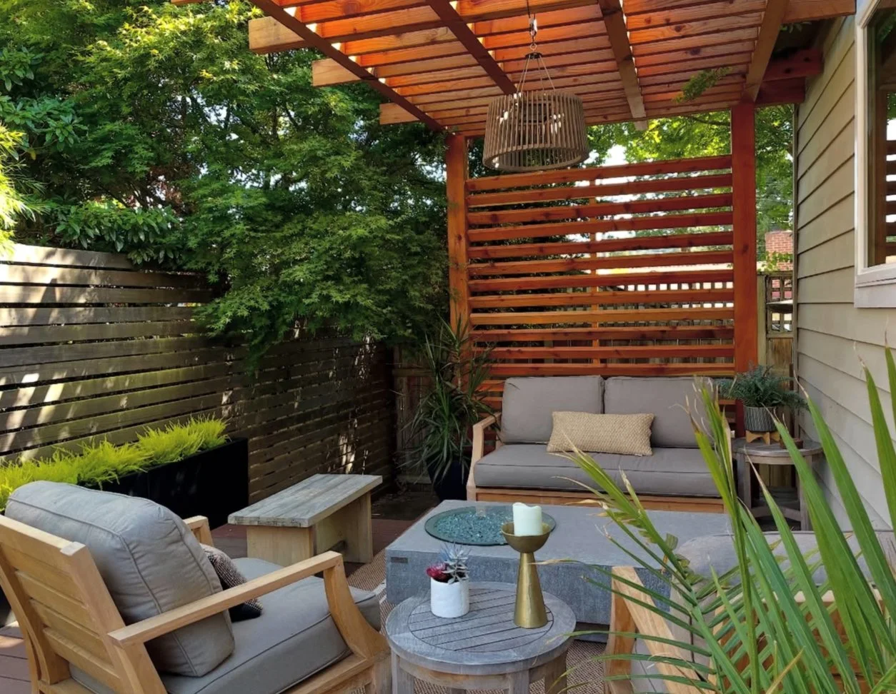 Outdoor patio with wooden furniture, cushioned seats, greenery, and a wooden slatted privacy fence under a wooden pergola with hanging light fixture.