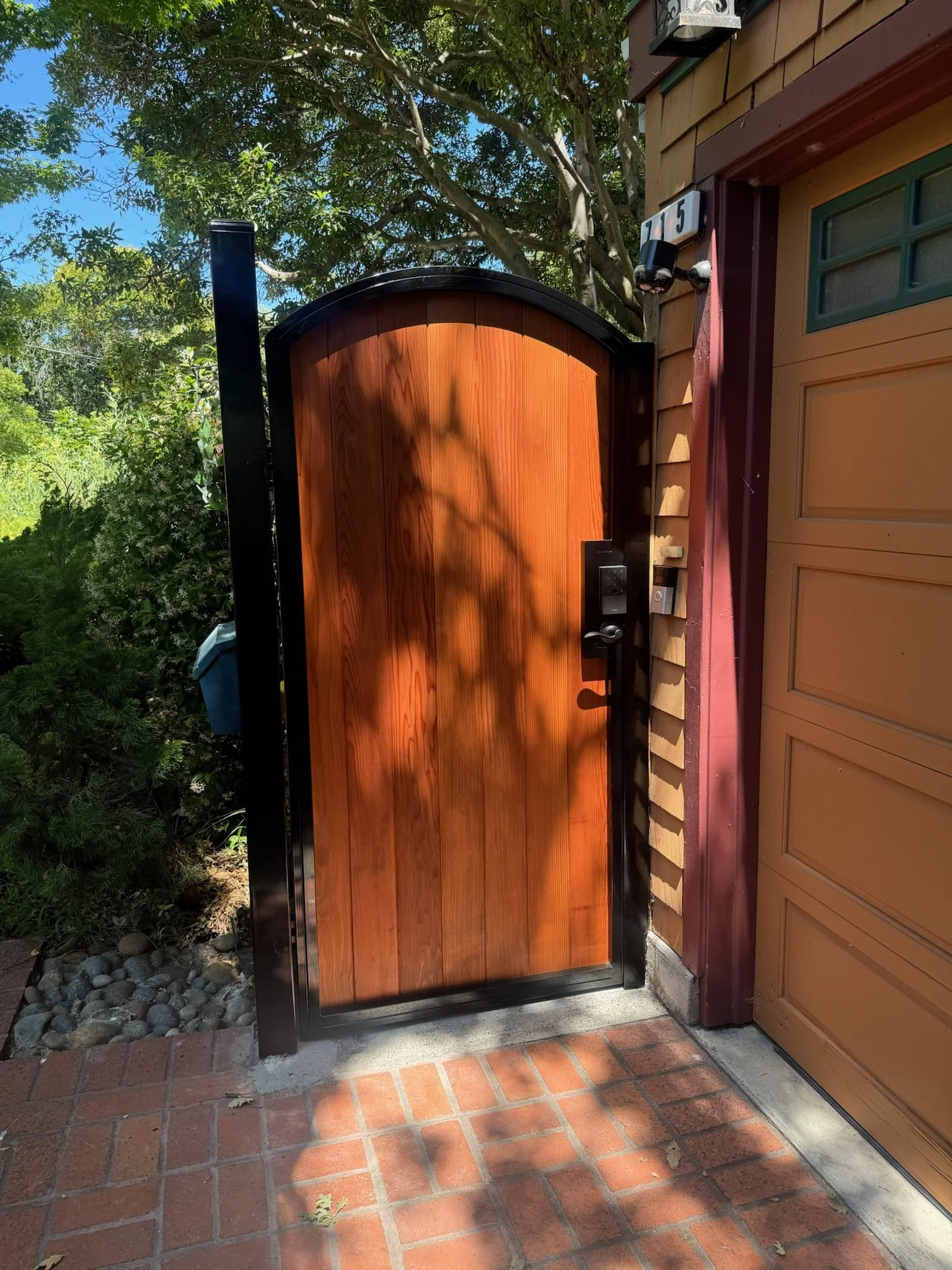A wooden gate with a black frame and a house number 15 on the wall beside it. The gate is in front of a garage door and surrounded by greenery and trees, with sunlight casting shadows.