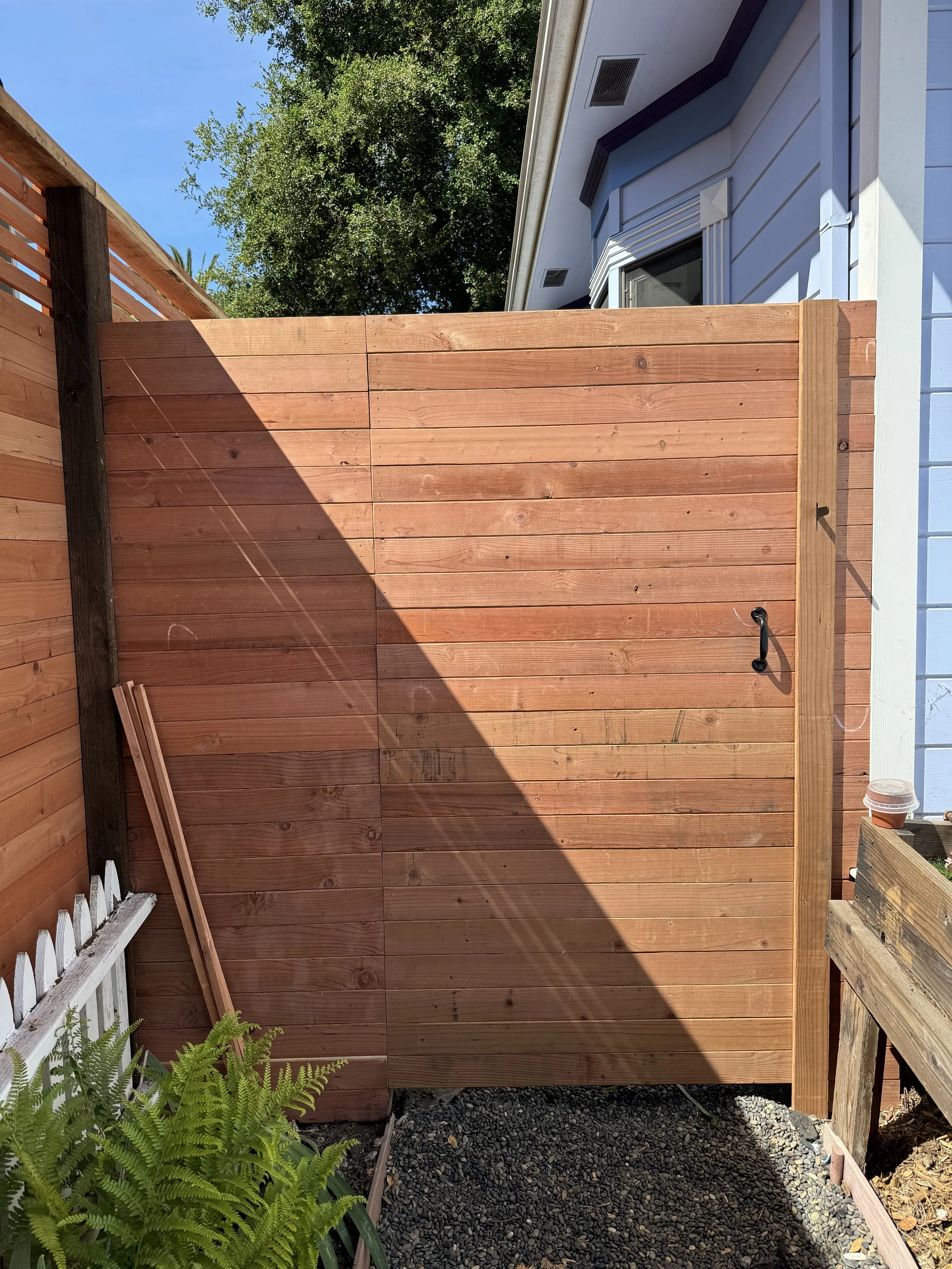 New wooden privacy fence with horizontal planks installed next to a house, with a black handle on the fence door, surrounded by greenery and a blue sky.