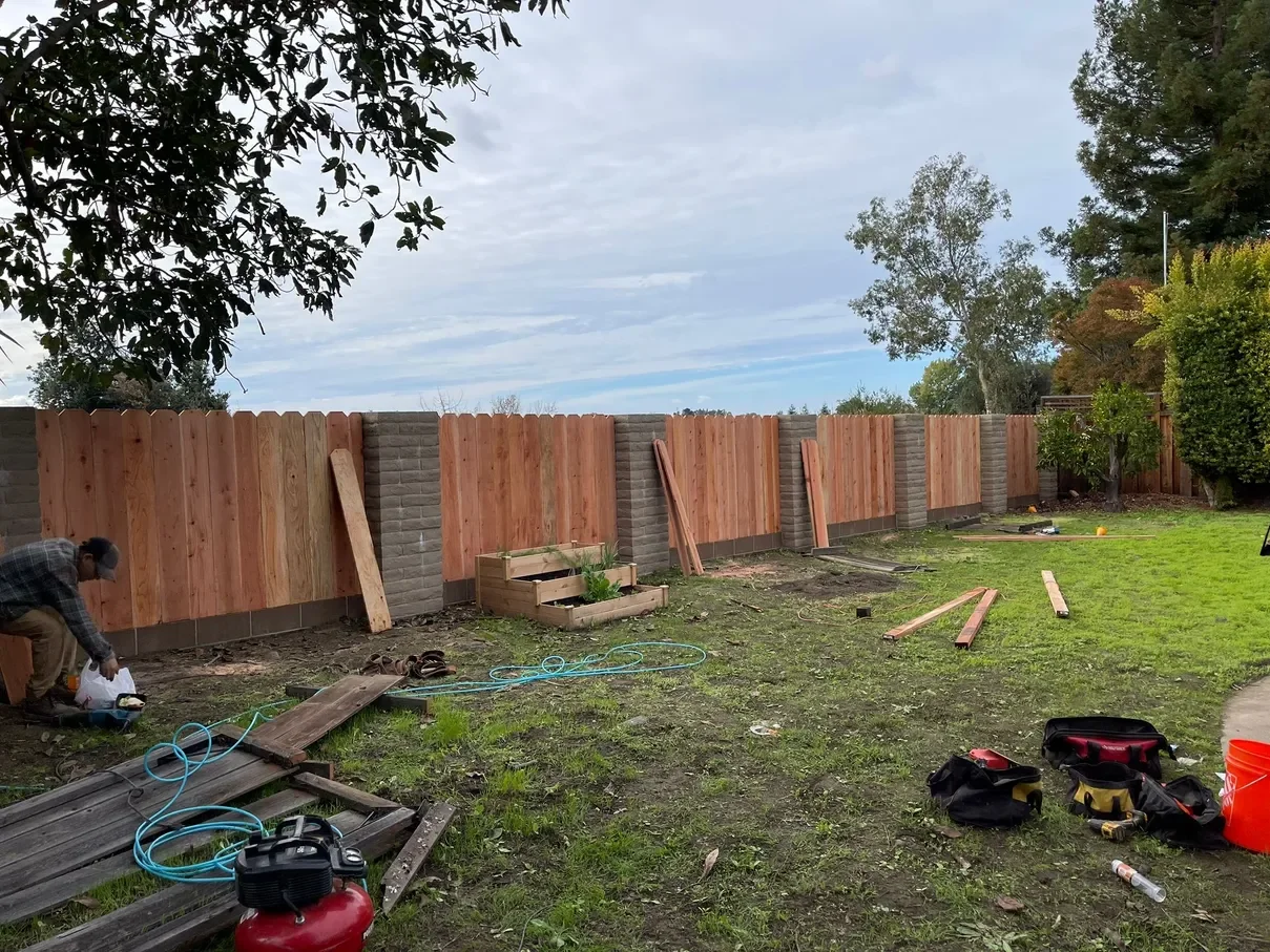 A backyard with a new wooden fence being built, construction tools and materials scattered around, and a worker on the left working on the project.