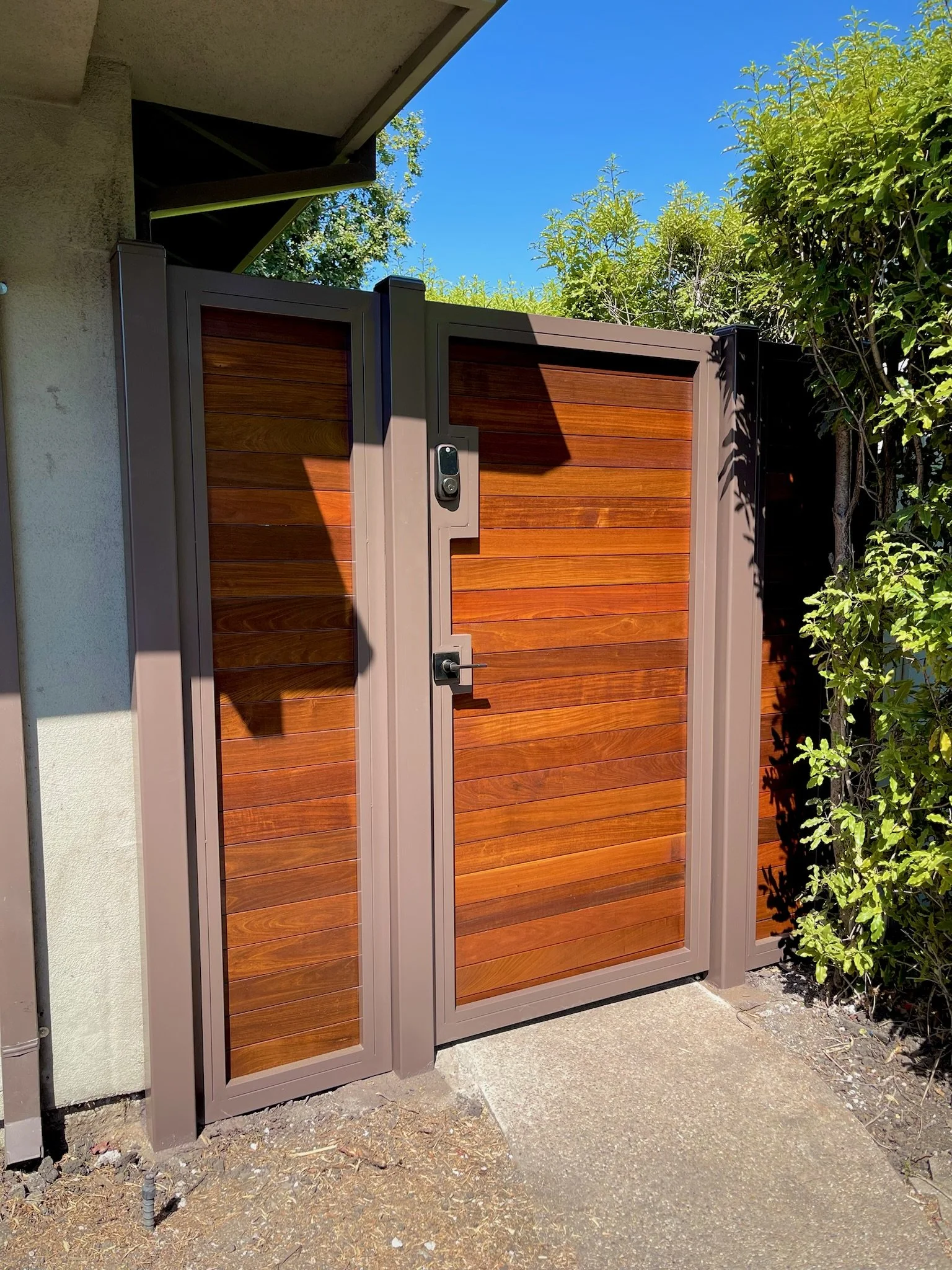 A modern outdoor gate with wood panel design and electronic access, set against a background of greenery and a bright blue sky.