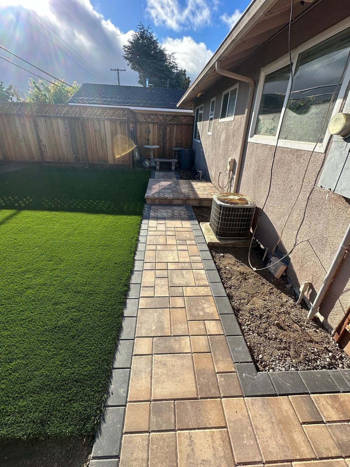 Backyard with a partially finished stone walkway, a small patio area, a green lawn, a wooden fence, and the side of a house with an air conditioning unit.