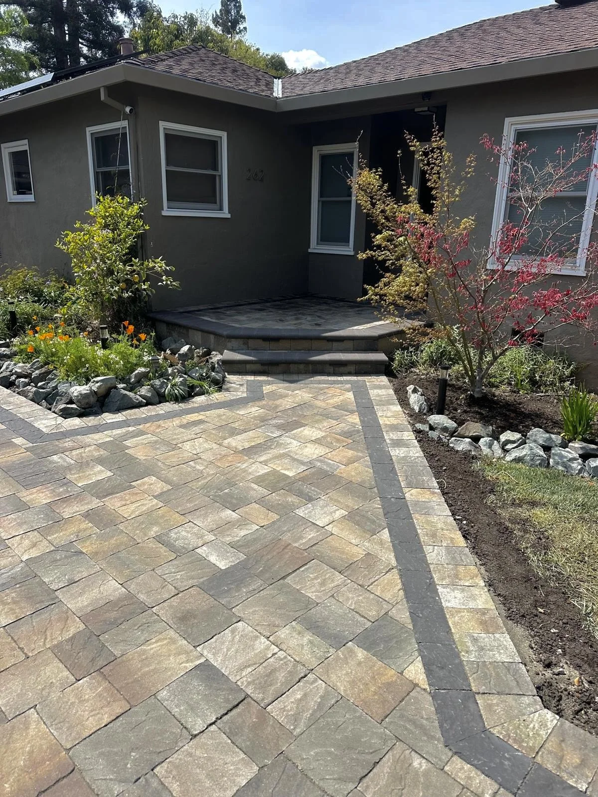 Front entry of a house with a paved walkway, small steps leading to the door, and landscaped garden beds with trees and flowers.