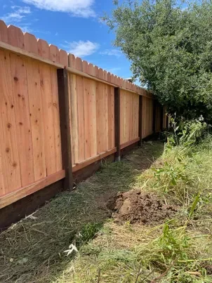 Wooden fence with support posts next to a grassy area and a tree under a blue sky.
