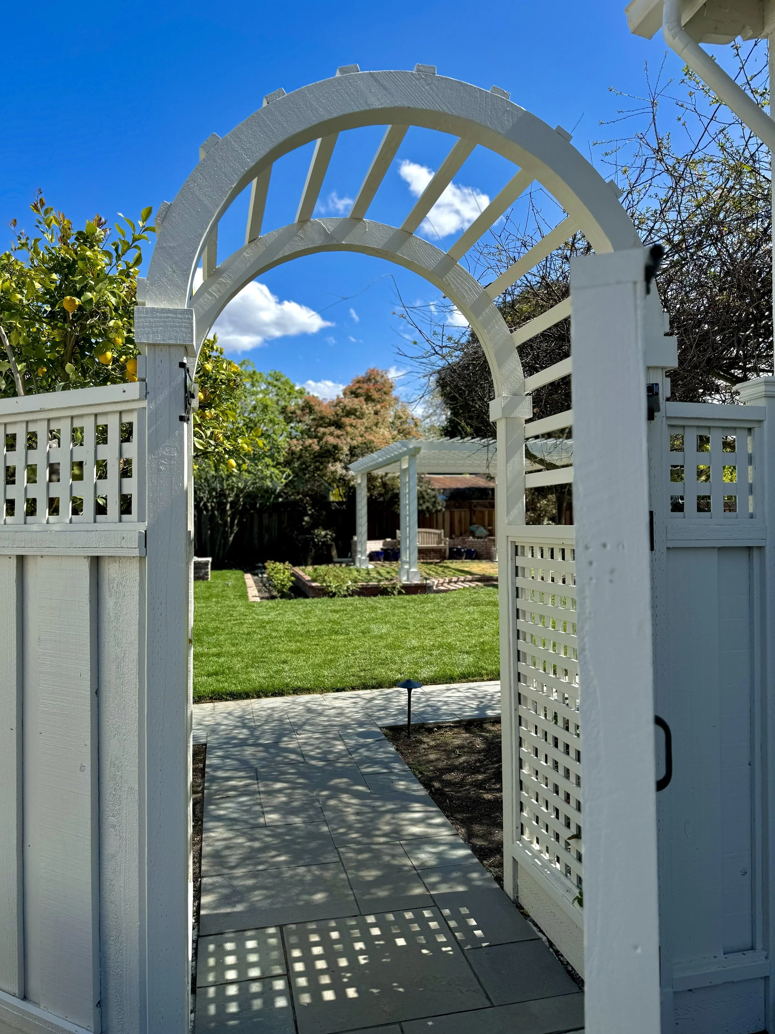 A white garden gate with a decorative archway opens to a well-maintained backyard with green grass, a paved walkway, a small garden bed, and a lattice fence, under a blue sky with scattered clouds.
