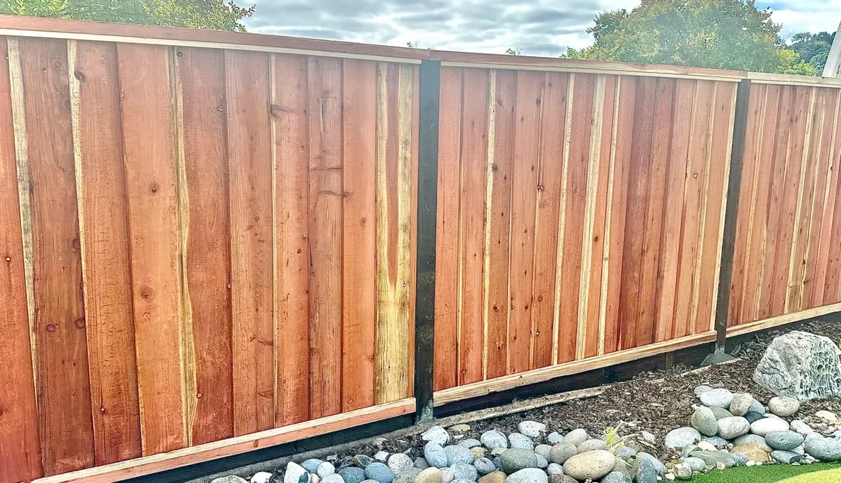 A wooden privacy fence with vertical planks and black metal posts, installed on a gravel bed with decorative rocks, under a cloudy sky.