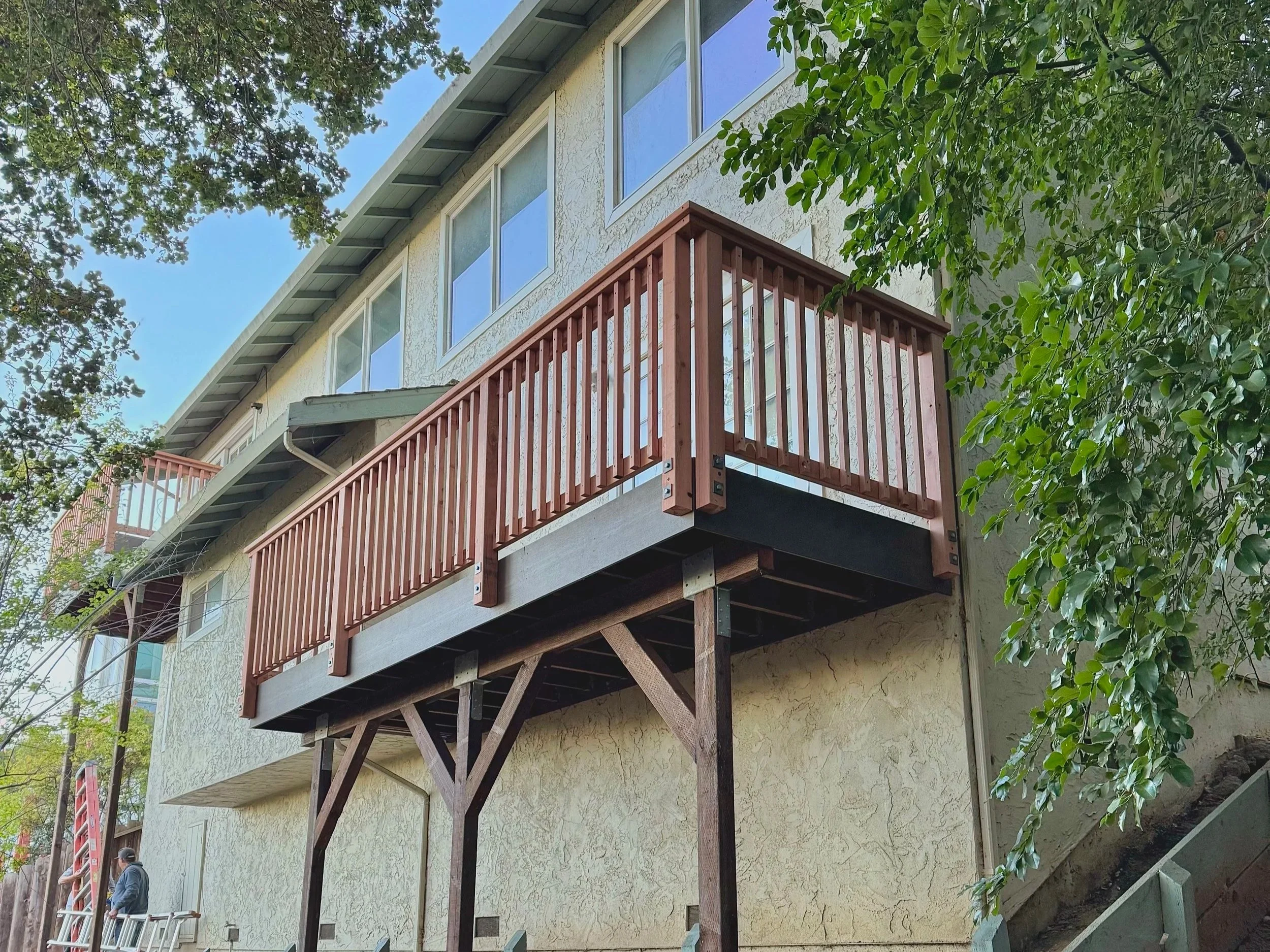 A newly built wooden balcony with railing extending from the second story of a house, supported by wooden beams, with trees and two people standing nearby.