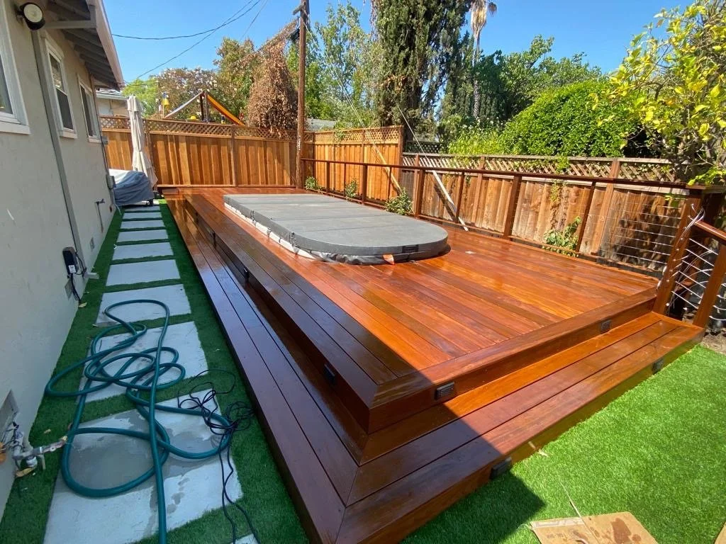 Backyard with a newly installed wooden deck with a hot tub on top, adjacent to a house with a concrete pathway and garden hose, surrounded by a wooden fence and trees.