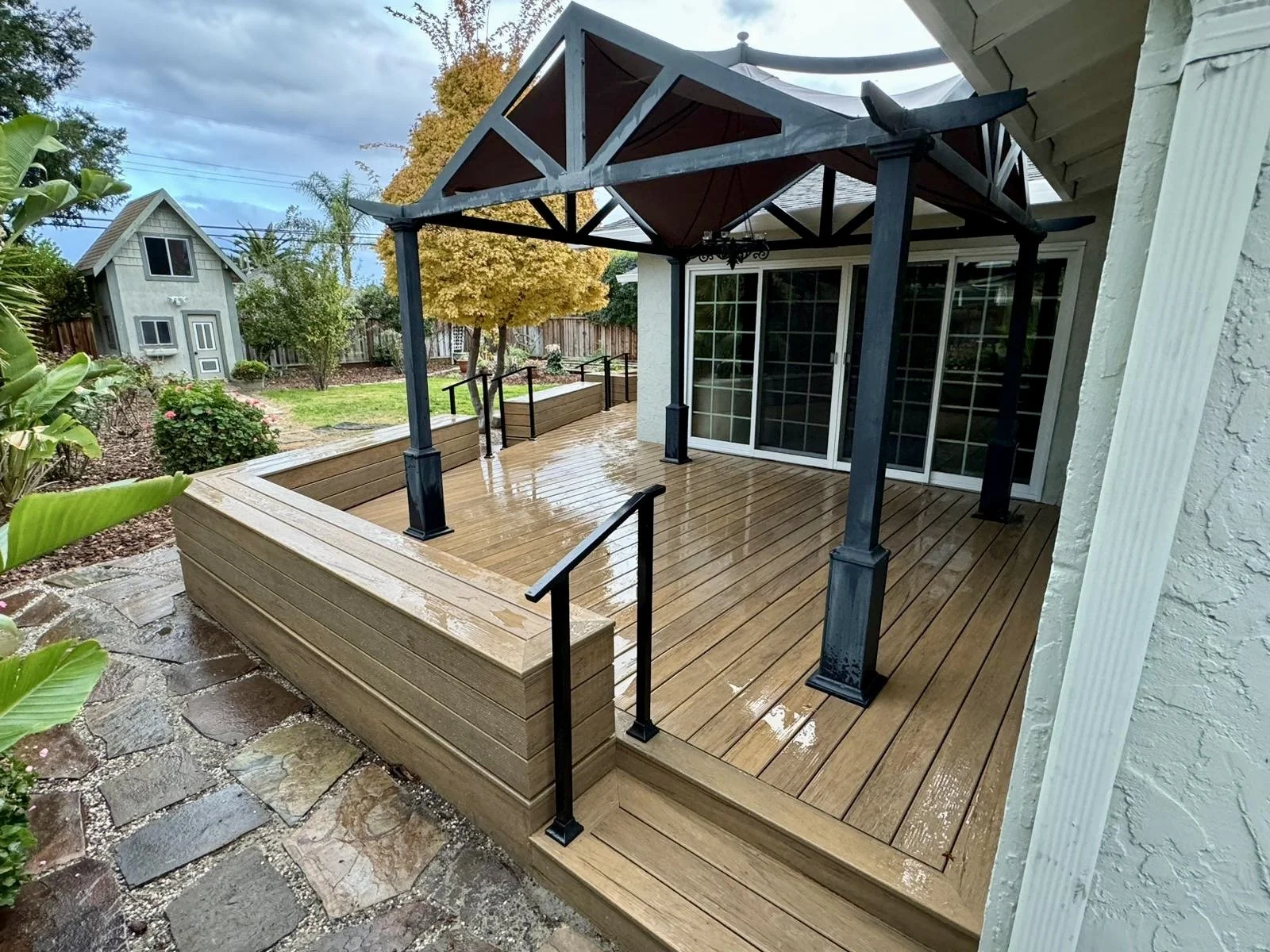 A wooden deck with a small covered porch attached to a house, rain droplets on the surface, overlooking a backyard with greenery, trees, and a small shed.