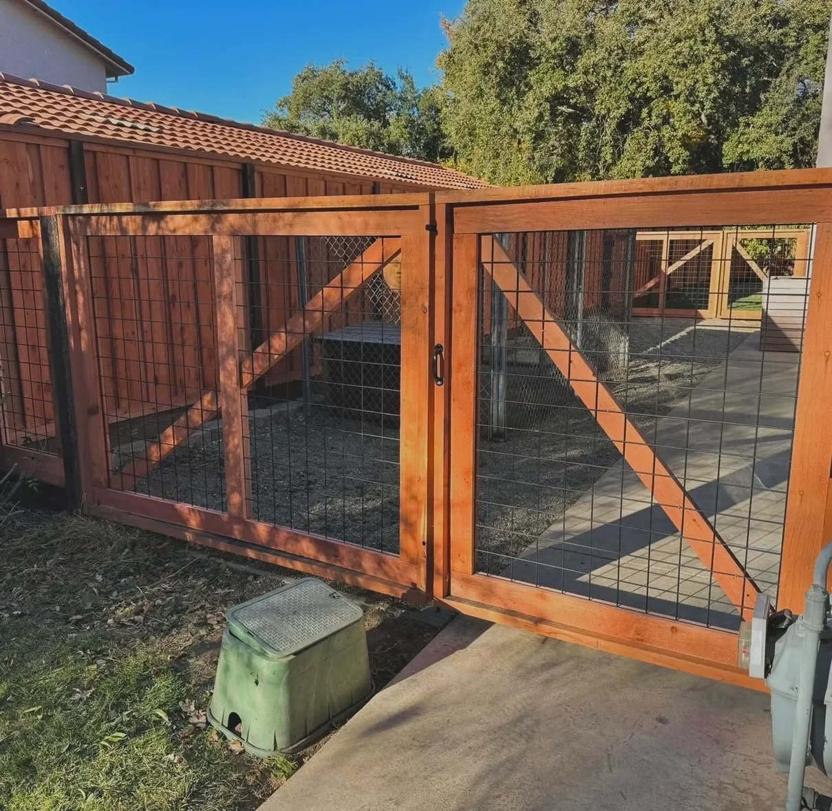 Newly installed wooden and wire mesh dog fence gate with handle, situated on concrete and dirt, with a green compost bin nearby, in a backyard with trees and a neighboring house with a tiled roof.