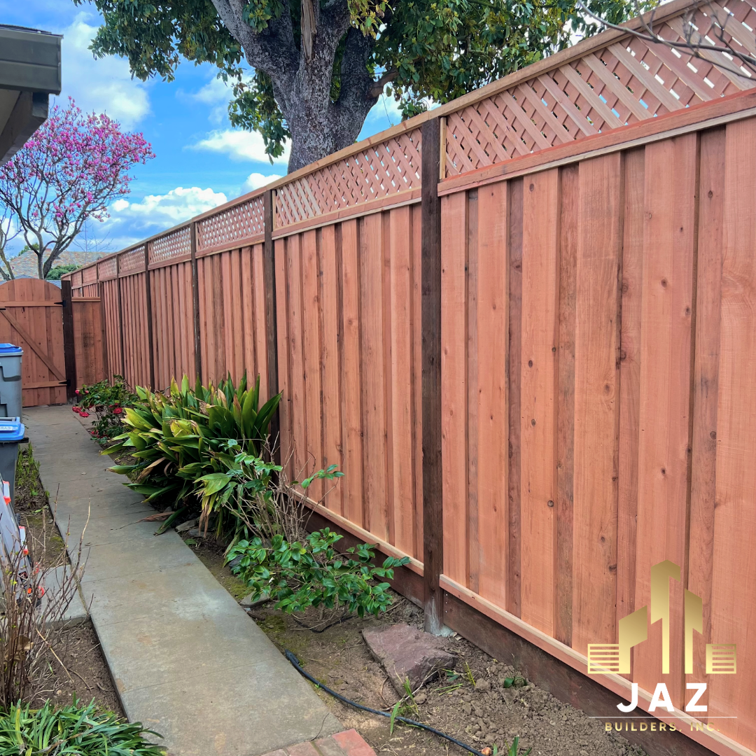 New wooden privacy fence installed along a garden pathway, with green plants and a large tree with dark bark behind it, under a blue sky with clouds.