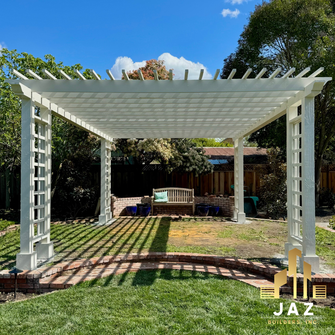 White pergola in a backyard with a brick patio, green grass, trees, and a wooden bench in the background