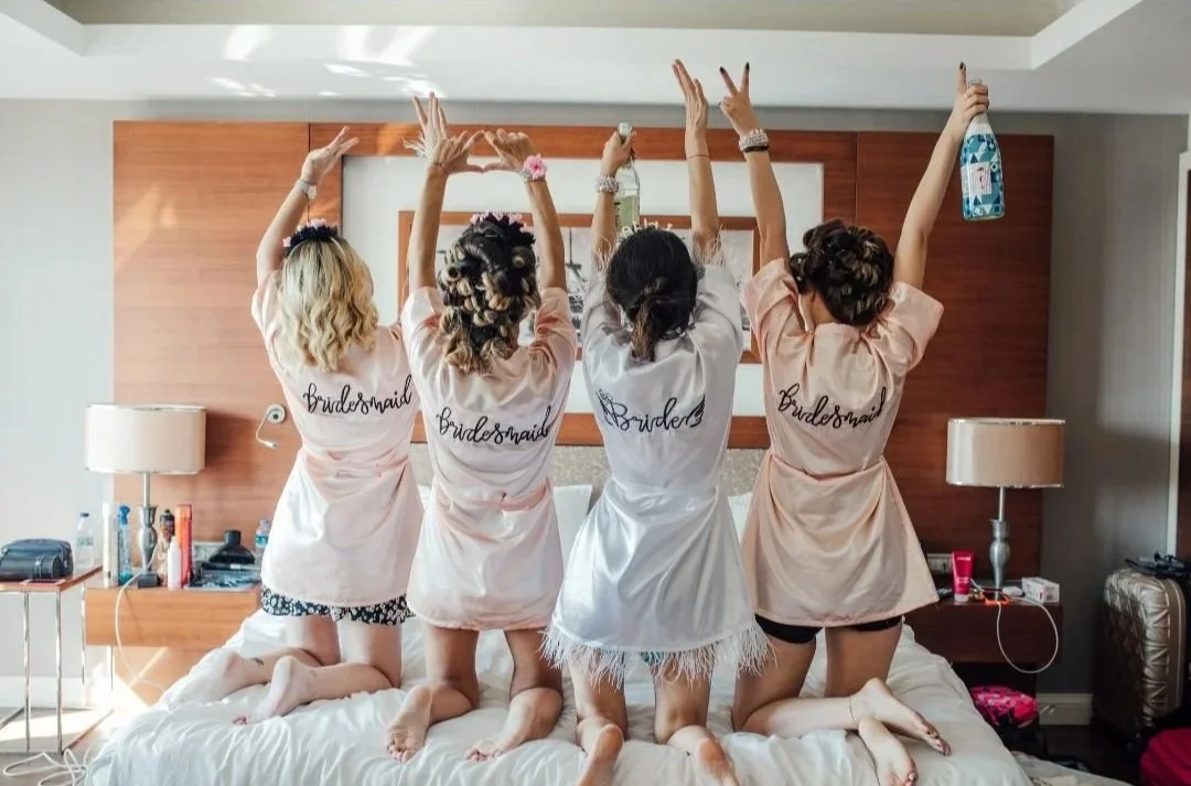 Four women in pajamas with 'Bridesmaid' text on the back, kneeling on a bed with their arms raised, back to the camera, in a hotel room.