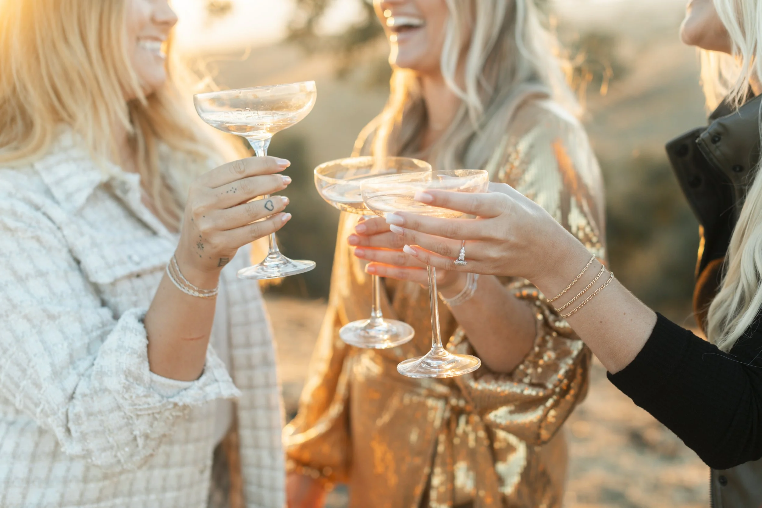Three women celebrating with champagne cocktails in coupe glasses, smiling and raising glasses outdoors at sunset.